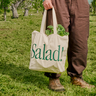 Close-up of person holding Salad! Collection Tote in a field filled with greens.