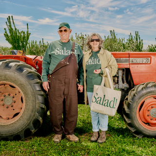 Couple holding hands and smiling wearing Salad! Collection apparel and accessories in front of a tractor.